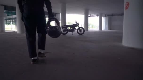 Байкер, идущий через парковку к мотоциклу. Мотоциклист и винтажный мотоцикл 1970-х годов. Сзади вид на городскую жизнь . — стоковое видео