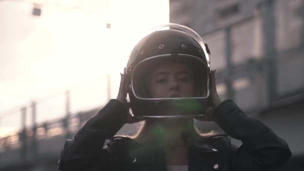 Close-up de jovem motociclista mulher decolando capacete retro ao ar livre ao pôr do sol. Menina motociclista em couro jaqueta vestir para viagem. Vista frontal, slow mo — Vídeo de Stock