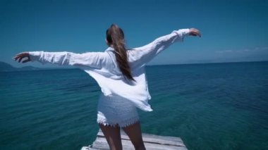 Beyaz gömlekli ve beyaz şortla turist kadın iskele boyunca gelen manzaranın tadını çıkarıyor ve ellerini kaldırıyor, deniz havasının ılık taze esintisinde kalıyor.