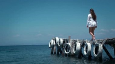 Beyaz gömlekli ve beyaz şortla turist kadın iskele boyunca gelen manzaranın tadını çıkarıyor ve ellerini kaldırıyor, deniz havasının ılık taze esintisinde kalıyor.