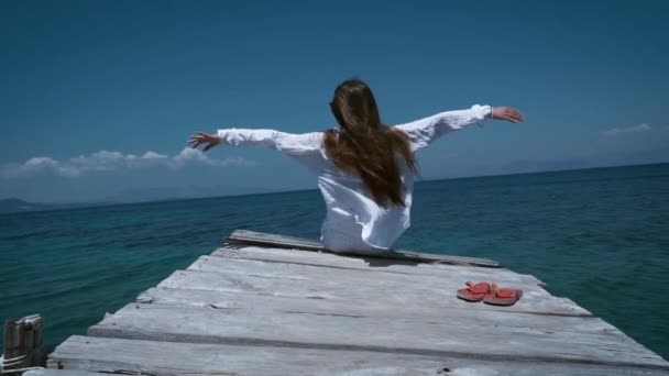 Retrato de una mujer sentada en un muelle de madera y levanta las manos mirando al mar, sintiendo libertad, sobre el fondo del agua azul marino y el cielo. Concepto: libertad, hermosa vista al mar — Vídeo de stock