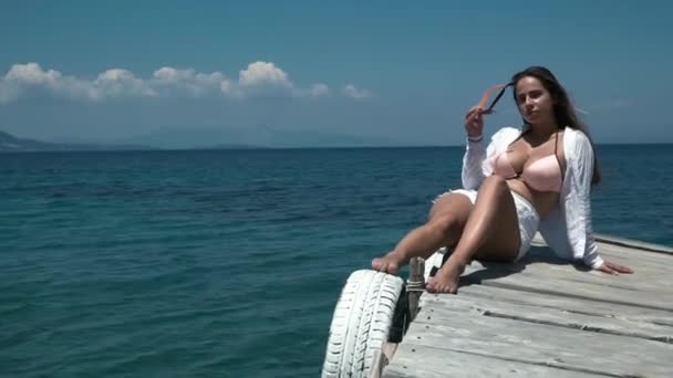 Mujer turista en camisa blanca y pantalones cortos blancos disfrutar de la vista, se sienta en el muelle y se quita las gafas de sol, permaneciendo en la cálida brisa fresca del aire del mar . — Vídeos de Stock