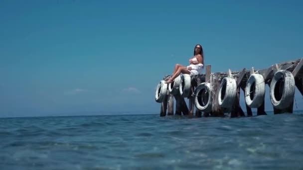 Tourist woman in white shirt and white shorts enjoy the view, sits on the dock, staying on the warm fresh breeze of the sea air. — Stock Video