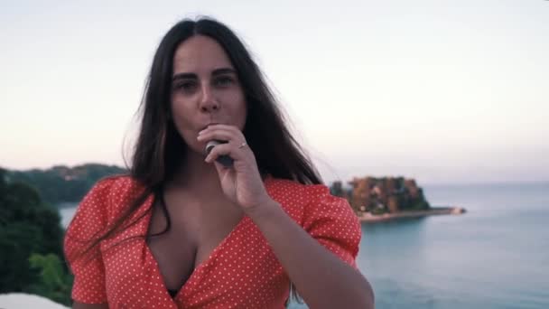 La chica fumando con dispositivo calentador de tabaco en vestido rojo, fondo de mar — Vídeos de Stock