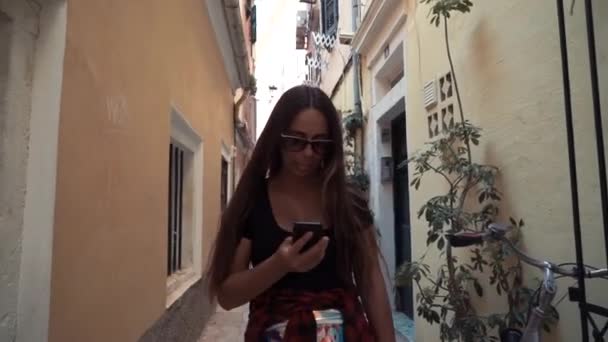 Alegre turista femenina en gafas de sol instalando navegador en el dispositivo telefónico moderno caminando por las calles en el entorno urbano.viajero positivo paseando — Vídeos de Stock