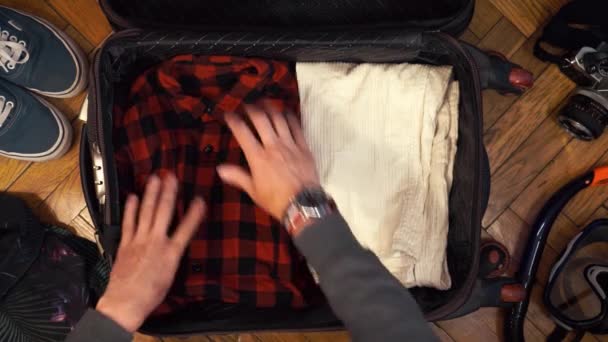 As mãos do homem estão a fazer malas para uma viagem. O homem sonha com viajar. Roupas de verão, óculos de sol e câmera. Vista superior — Vídeo de Stock