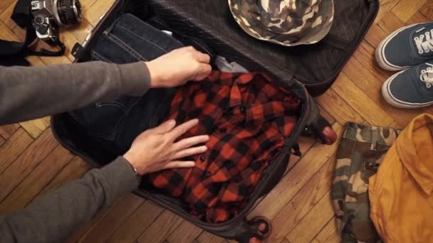 Joven con los brazos fuertes empaca su maleta llena de artículos artesanales varoniles como vaqueros de orillo, camisa de franela, frasco de acero inoxidable y otros accesorios hipster. Vista superior — Vídeos de Stock