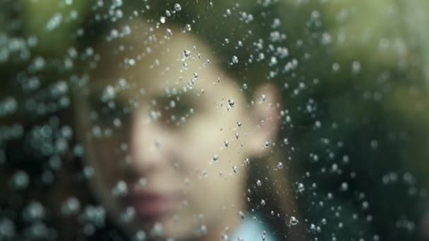 Jonge aantrekkelijke vrouw zit bij het raam. het regent buiten het raam. Portret van een mooi meisje dat naar de camera kijkt. grote druppels glijden over het glas. — Stockvideo