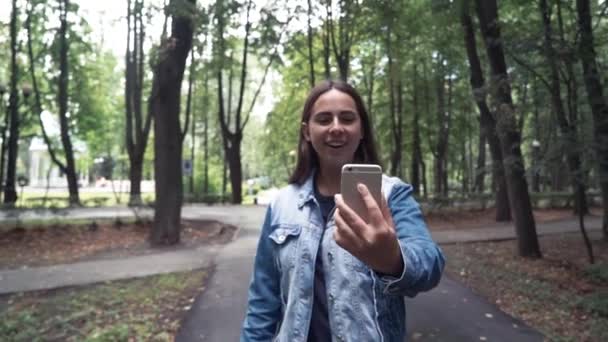 Молодая красивая женщина звонит по телефону в Жан Jacketwalking в летнем парке. видеовызов — стоковое видео