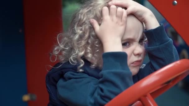 O menino se senta em um carro de madeira no Parque Infantil e brinca. O menino está com raiva e infeliz. A câmera segue uma criança sentada em um carro de brinquedo . — Vídeo de Stock
