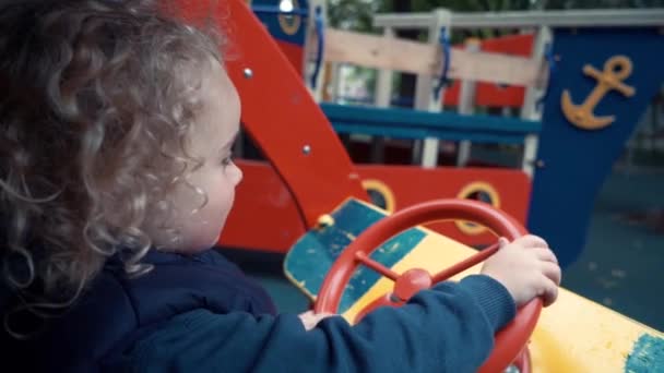 Il ragazzo è seduto in una macchina di legno sul campo da giuoco e gioca. La fotocamera monitora il bambino seduto in un'auto giocattolo . — Video Stock