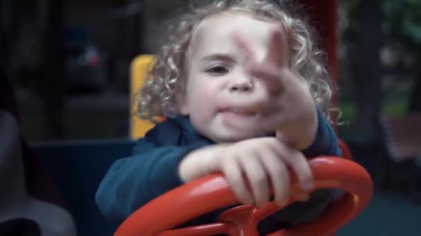 Pojken sitter i en träbil på lekplatsen och leker. Pojken är arg och olycklig. Kameran följer ett barn som sitter i en leksaksbil. — Stockvideo