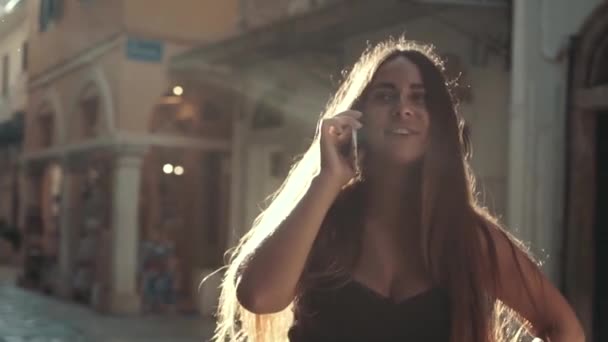 Young stylish woman walking at sunset and talking on phone. Joyful hipster girl using cellphone outdoors — Stock Video