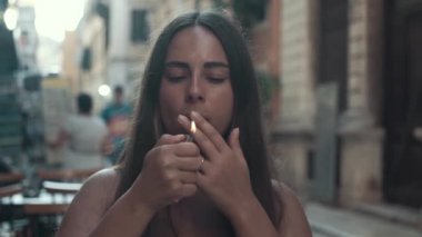 Genç bir kadın bir güneşli yaz sabahı bir kafede bir verandada otururken ince sigara içiyor. 4k, ağır çekim