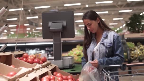 Молода брюнетка перевіряє червоні стиглі помідори, купує сезонні продукти в відділі свіжих овочів в супермаркеті . — стокове відео