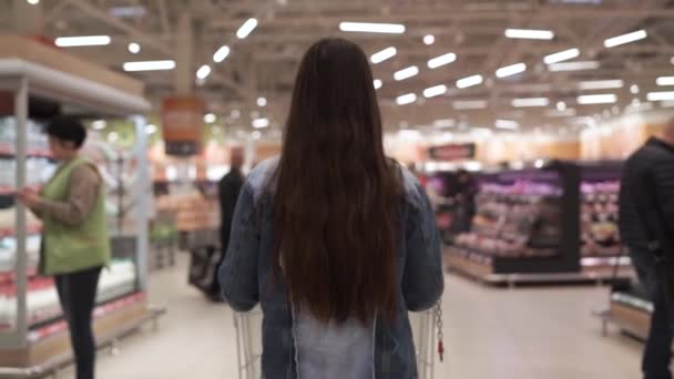 젊은 퍼티 갈색 머리는 대형 슈퍼마켓에서 걷고 음식을 사러 가는 쇼핑 트롤리를 밀어 내고 제품으로 선반을 둘러보고 있습니다. 사람과 상점 개념. 팔로우 샷 — 비디오