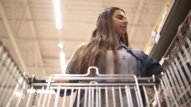 Rahat giysiler içinde Pretty kadın içinde gıda ile bakkal direksiyon alışveriş arabası yürüyor ve ürünleri ile raflarda etrafa bakıyor. Kadın ve dükkanlar kavramı.