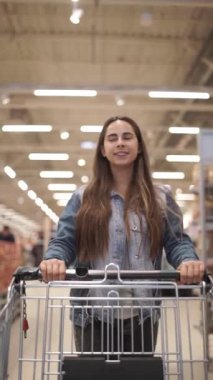 Rahat giysiler içinde Pretty kadın içinde gıda ile bakkal direksiyon alışveriş arabası yürüyor ve ürünleri ile raflarda etrafa bakıyor. Dikey video 9 :16