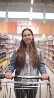 Rahat giysiler içinde Pretty kadın içinde gıda ile bakkal direksiyon alışveriş arabası yürüyor ve ürünleri ile raflarda etrafa bakıyor. Dikey video 9 :16