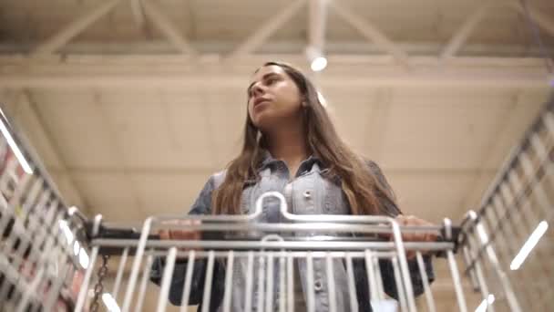 Mujer bonita en ropa casual está caminando en la tienda de comestibles volante carrito de la compra con comida dentro de ella y mirando a su alrededor en los estantes con productos. Concepto de mujeres y tiendas . — Vídeo de stock