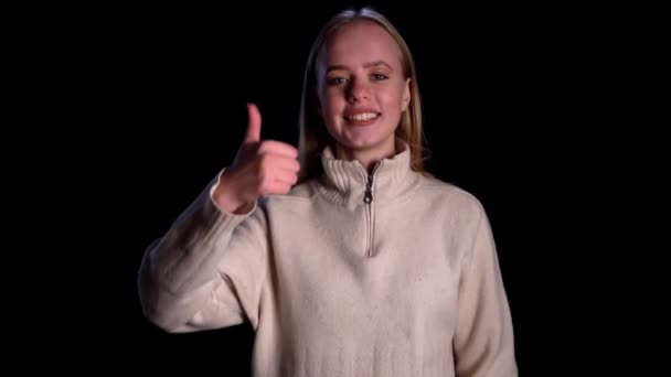 Boldog lány mutatja hüvelykujját a fekete háttér. Egy fiatal nő, aki kézjelként jelenik meg. Portré mosolygós lány mutatja hüvelykujját fel a stúdióban. — Stock videók