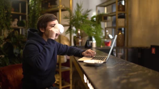 Hombre trabajando en un aptop en la cafetería. Joven freelancer está sentado en la cafetería con una taza de cofee en la mesa y escribiendo o buscando alguna información . — Vídeo de stock