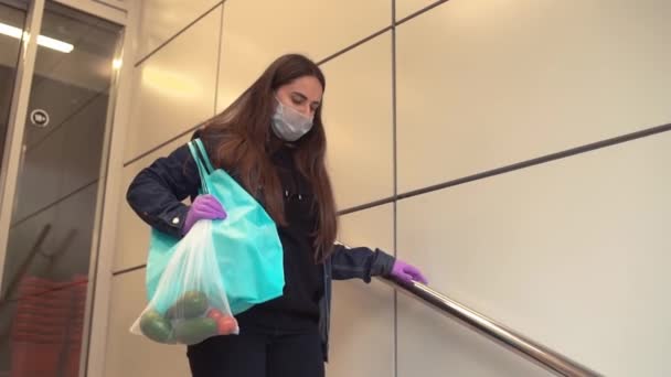 Доставка їжі молодою жінкою в захисну маску і рукавички несе сумку з продуктами з магазину. Концепція пандемії коронавірусу — стокове відео