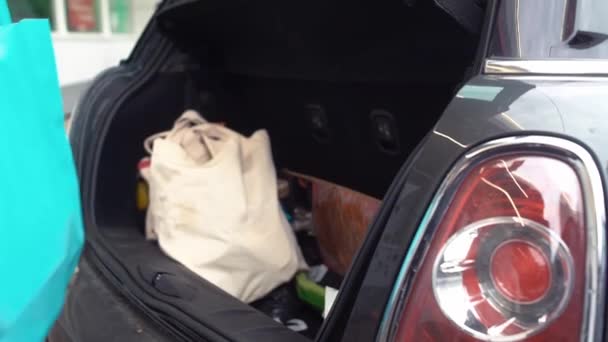 En kvinna i mask och handskar lägger shoppingväskor i bakluckan på en bil. Begreppet pandemiskt koronavirus — Stockvideo