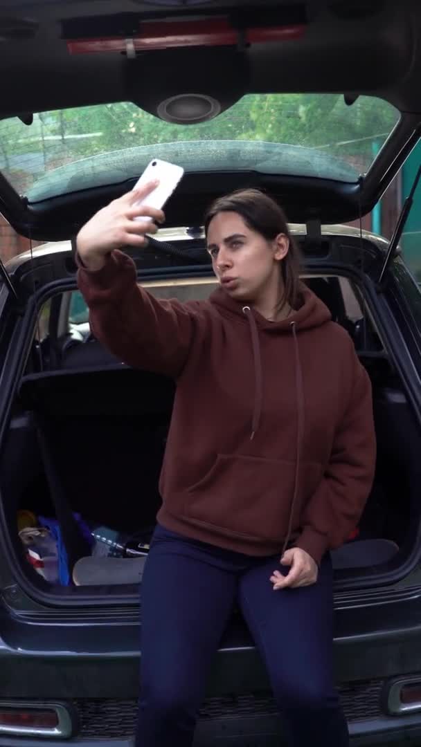 Sorglos aufgeregte junge Frau beim Selfie mit dem Smartphone im stilvollen Kofferraum. Positives Mädchen posiert für ein Selfie, macht lustige Gesichter und Mienen während eines sommerlichen Roadtrips. Vertikales Video — Stockvideo