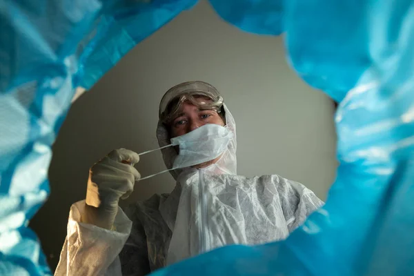 Fin de la victoria de cuarentena sobre el coronavirus. Vista desde el bote de basura un médico hombre se quita una máscara médica y guantes de látex y los tira. Papelera con equipo de protección personal desechable Imagen De Stock