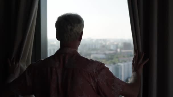 Young man silhouette opens curtains standing at large window against city buildings on summer day slow backside motion — Stock Video