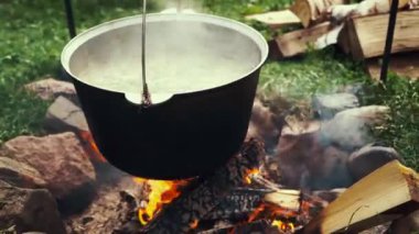 Siyah turist tenceresi karanlık ormanda yanan kamp ateşinin üzerinde asılı duruyor. Vahşi doğa yürüyüşünde yemek pişirmek. Sıcak çay ya da yiyecek kazanları ateşte ısıtılır. Açık hava seyahati. Kampta sıcak yemek kaynıyor.