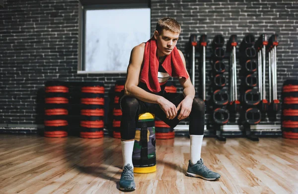 Full body view of handsome fit man relaxing after exercises with red towel on neck in the gym. Tired athletic male rest after hard workout with weight training with copy space for text, advertising