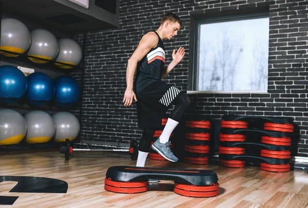 Side full body view of sporty athlete man doing step up jumps in health fitness club. The handsome male is exercising his legs on the stepper in the modern gym. Sport, lifestyle and people concept.