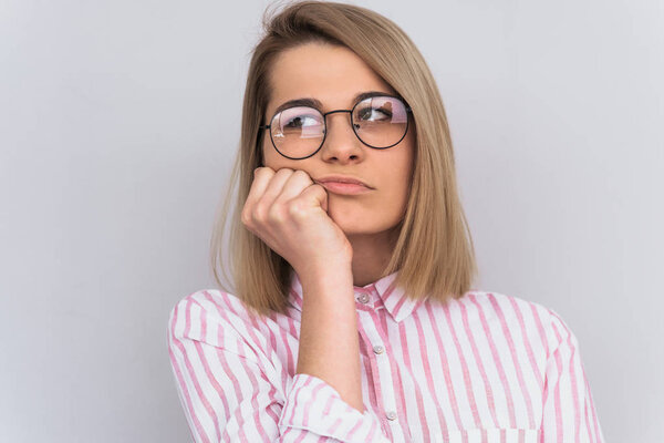 Studio portrait of sad blonde female student wearing pink shirt and round spectacles, sitting with unhappy boring expression on her face, resting chin on hand listening information. People, emotion 