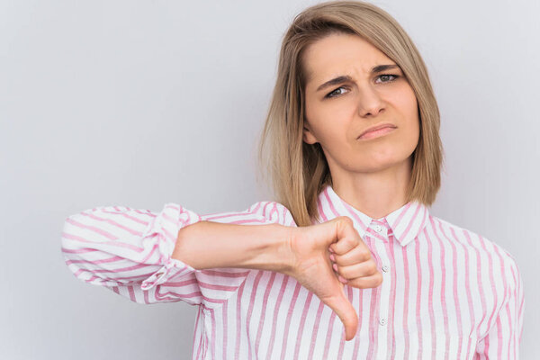 Closeup portrait of unhappy blonde female wears striped pink shirt looking at the camera with digusted expression, showing thumbs down. People, body language and emotion concept