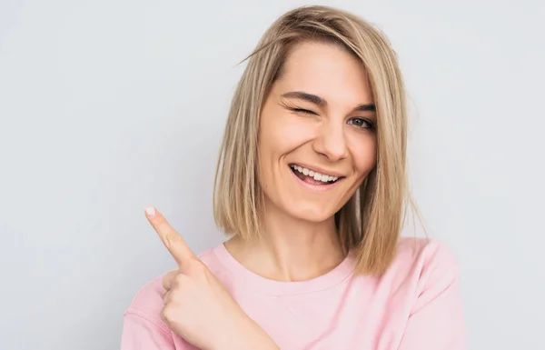 Closeup studio shot of happy blonde female wears pink sweater indicates with fore finger aside, blink with eye, demonstrates something at blank copy space for advertisement. People and emotion concept — Stock Image Closeup Studio Shot Happy Blonde Female Wears Pink Sweater Indicates Royalty Free Stock Photos
