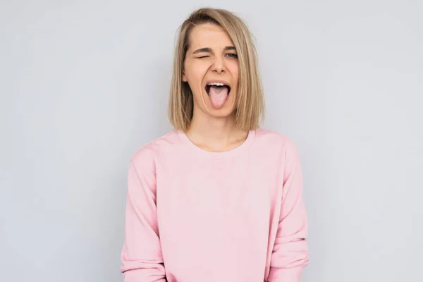 Portrait of disgusted pretty female sticking out tongue, expressing her disagreement to something. Beutiful funny young woman showing tongue at camera and blink with eye, posing over white wall.People — Stock Image Portrait Disgusted Pretty Female Sticking Out Tongue Expressing Her Disagreement Stock Image