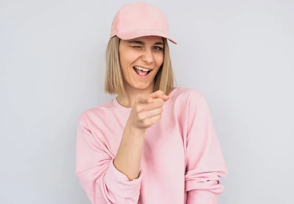 Portrait of blonde female blink with eye, wears pink clothes and cap, points at you, chooses someone or advertises product. Beautiful woman indicates at camera, isolated over white wall background — Stock Image Portrait Blonde Female Blink Eye Wears Pink Clothes Cap Points Stock Picture