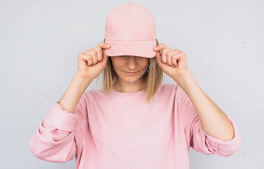 Closeup portrait of Caucasian blonde young female wearing pink sweater and cover her eyes with pink cap, protect from the sun rays, isolated on white studio background. Positive people emotion concept