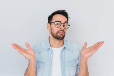 Portrait of uncertain young stylish stubble man with trendy round glasses wears demin blue shirt, shrugs shoulders being puzzled or confused. Caucasian unsure male make gestures doubtfully with hands. clipart