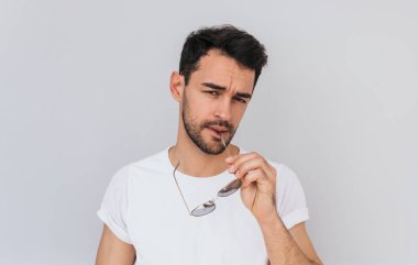 Seksi ciddi Genç sakallı erkek Casual beyaz T-shirt giymiş tutar güneş gözlüğü el ile beyaz beton zemin üzerinde izole kamera açık bakarak. İnsanlar, yaşam tarzı, moda kavramı