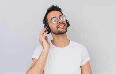Stüdyo yatay kapalı gözler ile favori müzik dinlerken kulaklık el ile holding beyaz tshirt ve mirros gözlük, mutlu genç erkek portresi beyaz arka plan üzerinde izole. Kopya alanı