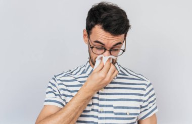 Closeup portrait of Caucasian sick male wearing striped shirt and glasses, caught cold and sneezing into tissue. Man have headache, virus against white background. Healthy, medicine concept. clipart