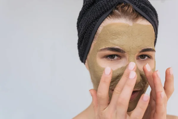Studio closeup portrait of young woman applying facial cosmetic green clay organic mask on her face, wears black towel on hair. Female taking care of her face skin, isolated over white wall. Healthy