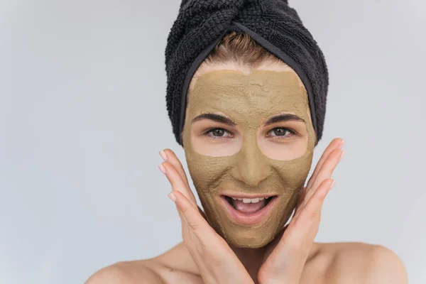 Studio portrait of happy young woman applying facial cosmetic green clay organic mask on her face, wears black towel on hair. Female taking care of her face skin, isolated over white wall. Healthy