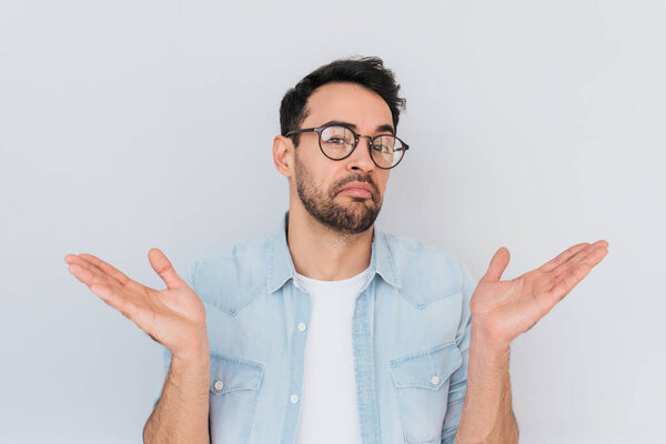 Portrait of uncertain young stylish stubble man with trendy round glasses wears demin blue shirt, shrugs shoulders being puzzled or confused. Caucasian unsure male make gestures doubtfully with hands.