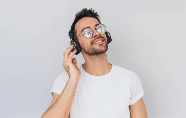 工作室水平画像的快乐年轻男性身穿白色 T恤和镜子眼镜 手持耳机与手 听最喜欢的音乐与闭上眼睛孤立的白色背景 复制空间 — 图库照片