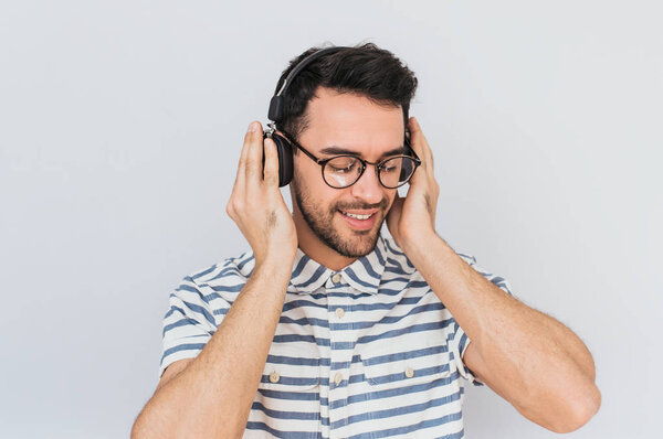 Horizontal shot of handsome happy young male wearing striped shirt and trendy glasses, with headphones on head, listening favorite music, isolated on white studio background.Copy space for advertising