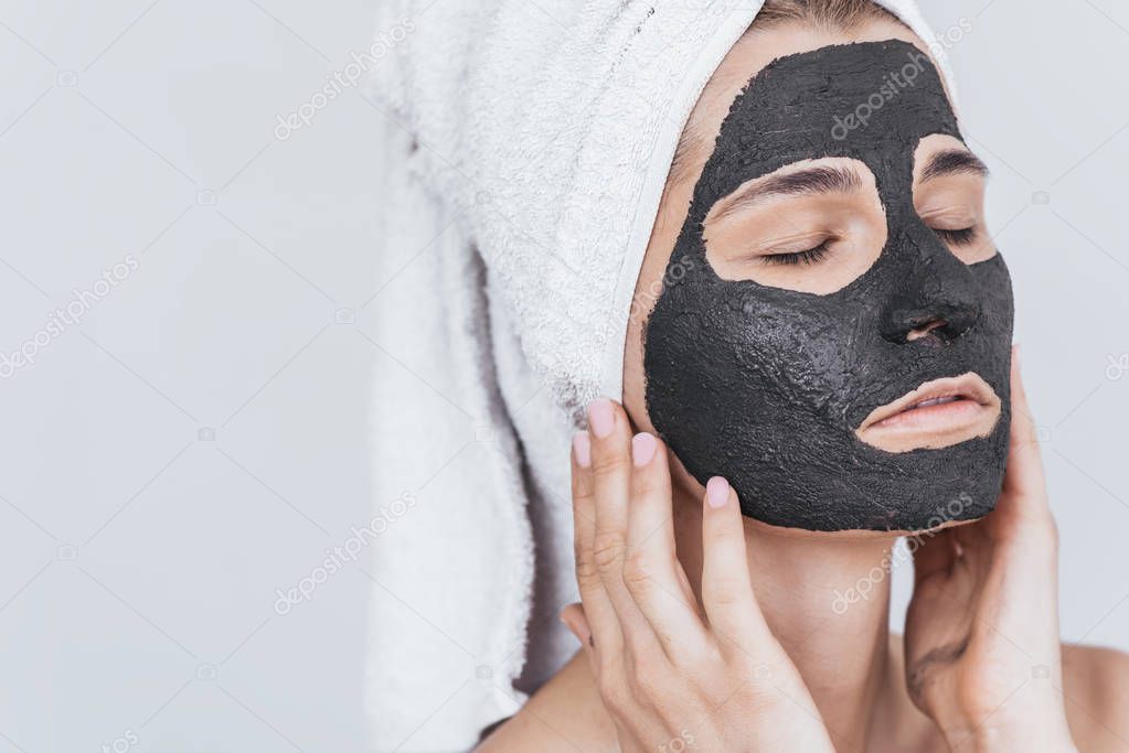 Closeup shot of young beautiful female applying black clay mask, doing beauty wellness treatment on her face skin. Horizontal shot of woman applying organic facial mask, with closed eyes on white